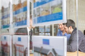 Ingatlaniroda kirakatán keresztül fotózott jelenet: egy fiatal pár és egy ingatlanügynök bent ülnek az irodában, asztalnál beszélgetnek, miközben a kirakatban eladó ingatlanok hirdetései láthatók.