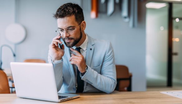 Ingatlanközvetítő öltönyben ül a laptopja előtt, és telefonál.