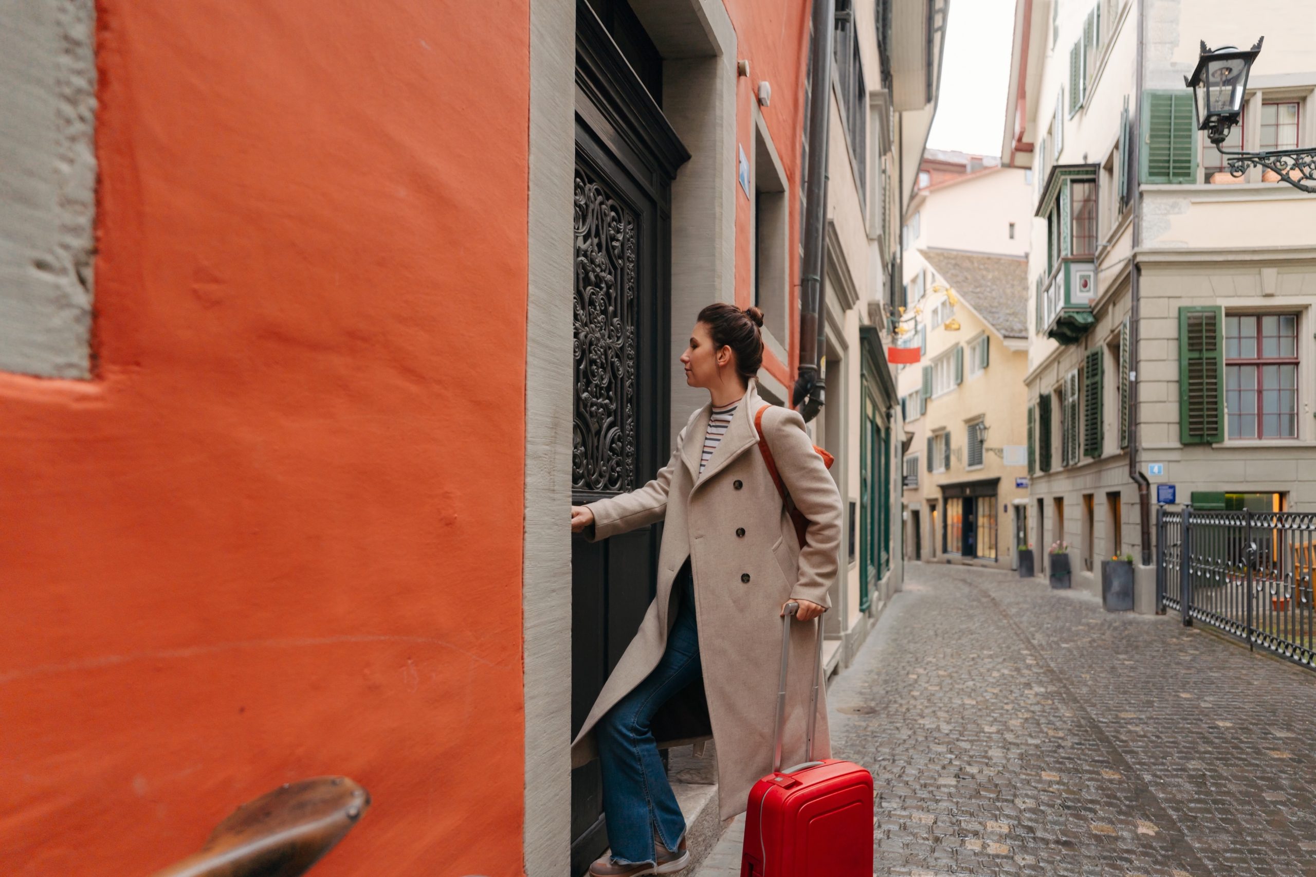 Albérlethez vagy rövid távú lakhatáshoz érkező nő egy régi városrész macskaköves utcájában: bőrönddel a kezében belép egy lakóépület kapuján. A kép a költözést, lakásbérlést, új otthonba érkezést és városi lakhatási helyzeteket illusztrálja.