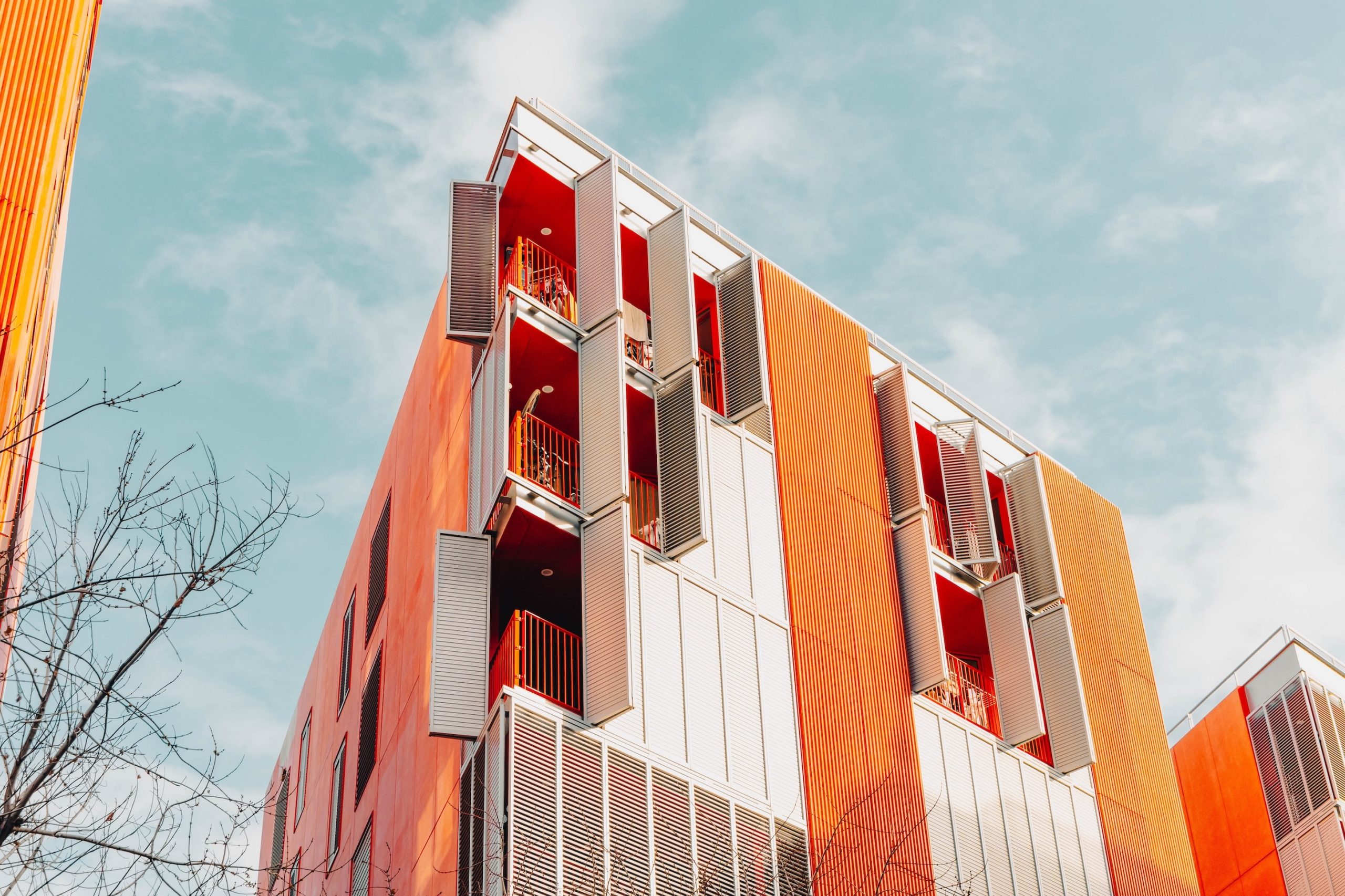 Modern, többemeletes társasház alulnézetből fotózva, narancssárga és világosszürke homlokzati elemekkel. Az épületen egymás fölött elhelyezkedő erkélyek láthatók, részben nyitható árnyékoló panelekkel és piros korlátokkal. A letisztult, kortárs építészeti stílusú lakóépület előtt egy lombtalan fa ága jelenik meg, a háttérben világoskék, enyhén felhős égbolttal.