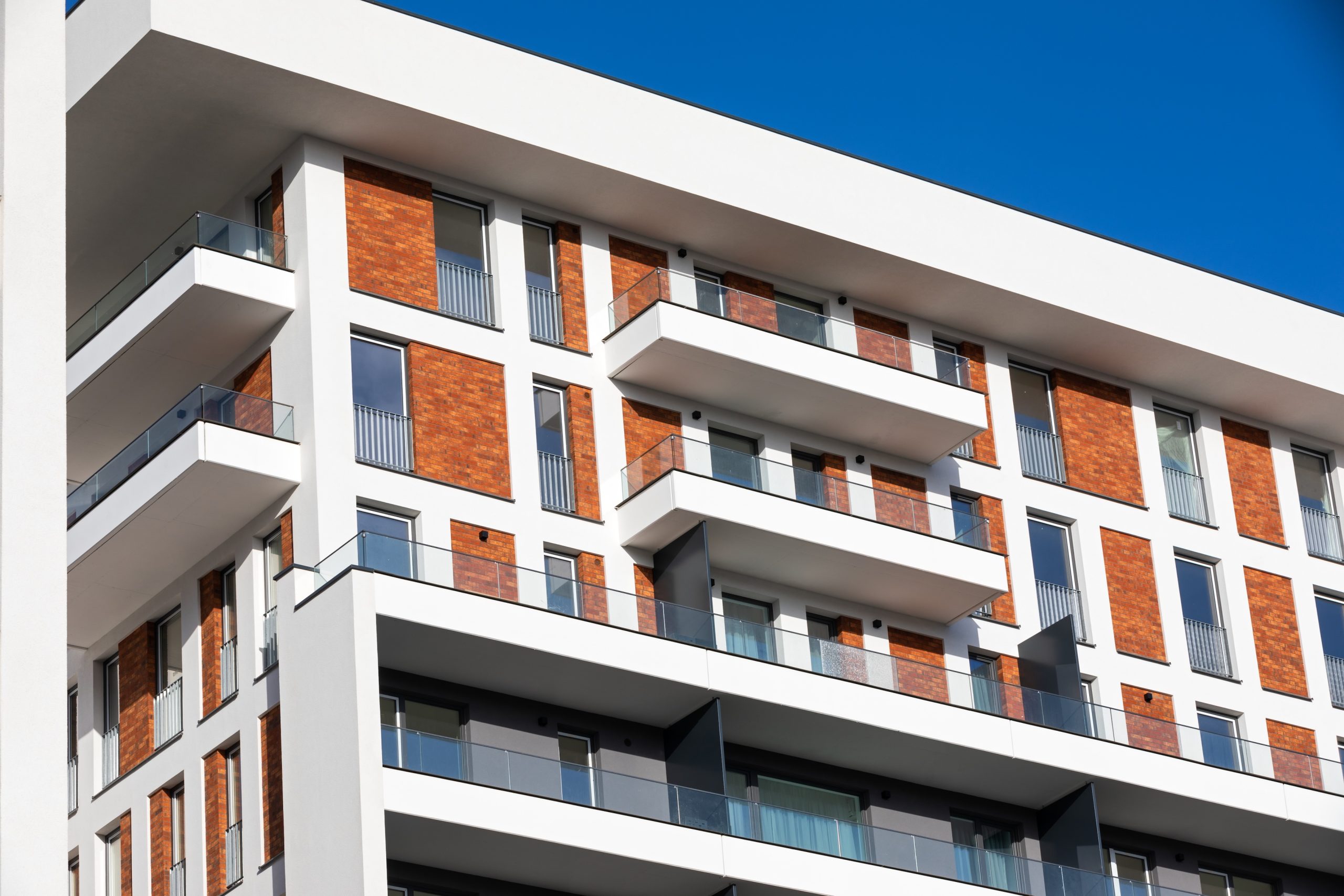 Modern, új építésű társasház erkélyes lakásokkal, fehér homlokzattal és téglabetétekkel, tiszta kék ég alatt.