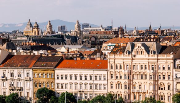 Budapest belvárosának panorámája historikus bérházakkal és díszes homlokzatú épületekkel. Az előtérben világos színű, többemeletes, klasszicista és eklektikus stílusú házak sorakoznak vörös cseréptetőkkel. A háttérben templomtornyok, kupolák és egyéb jellegzetes budapesti épületek emelkednek ki a város látképéből, távolabb pedig hegyek láthatók. A kép a főváros sűrűn beépített, történelmi városszövetét mutatja napsütéses időben.