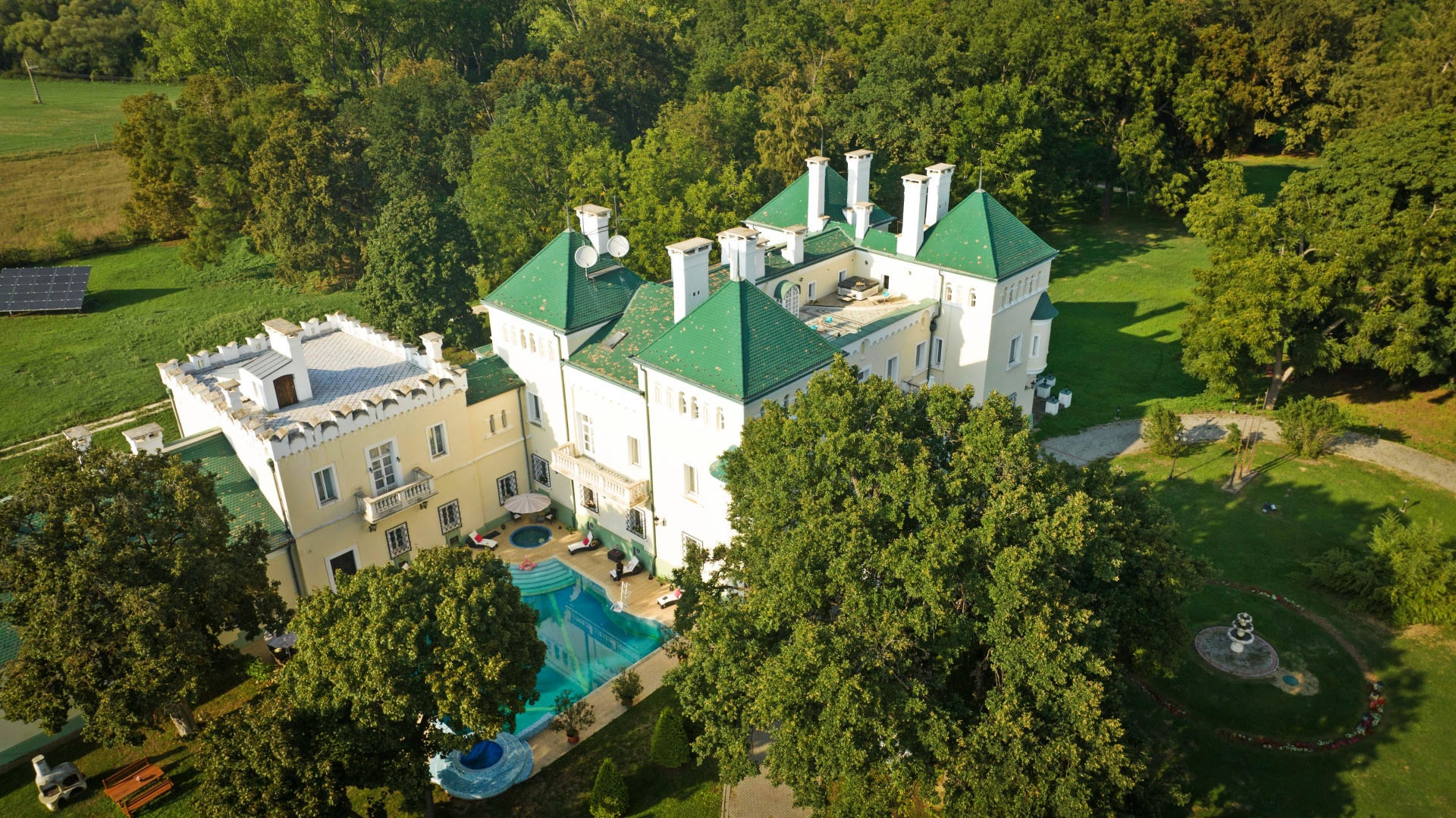Légi felvétel egy kastélyszerű, nagy méretű villáról zöld parkos környezetben. Az épület világos színű falakkal és zöld tetőkkel rendelkezik, több toronnyal és kéménnyel. Az udvaron egy medence és pihenőrész látható, körülötte gondozott kert, fák és sétányok, a háttérben erdős terület terül el.