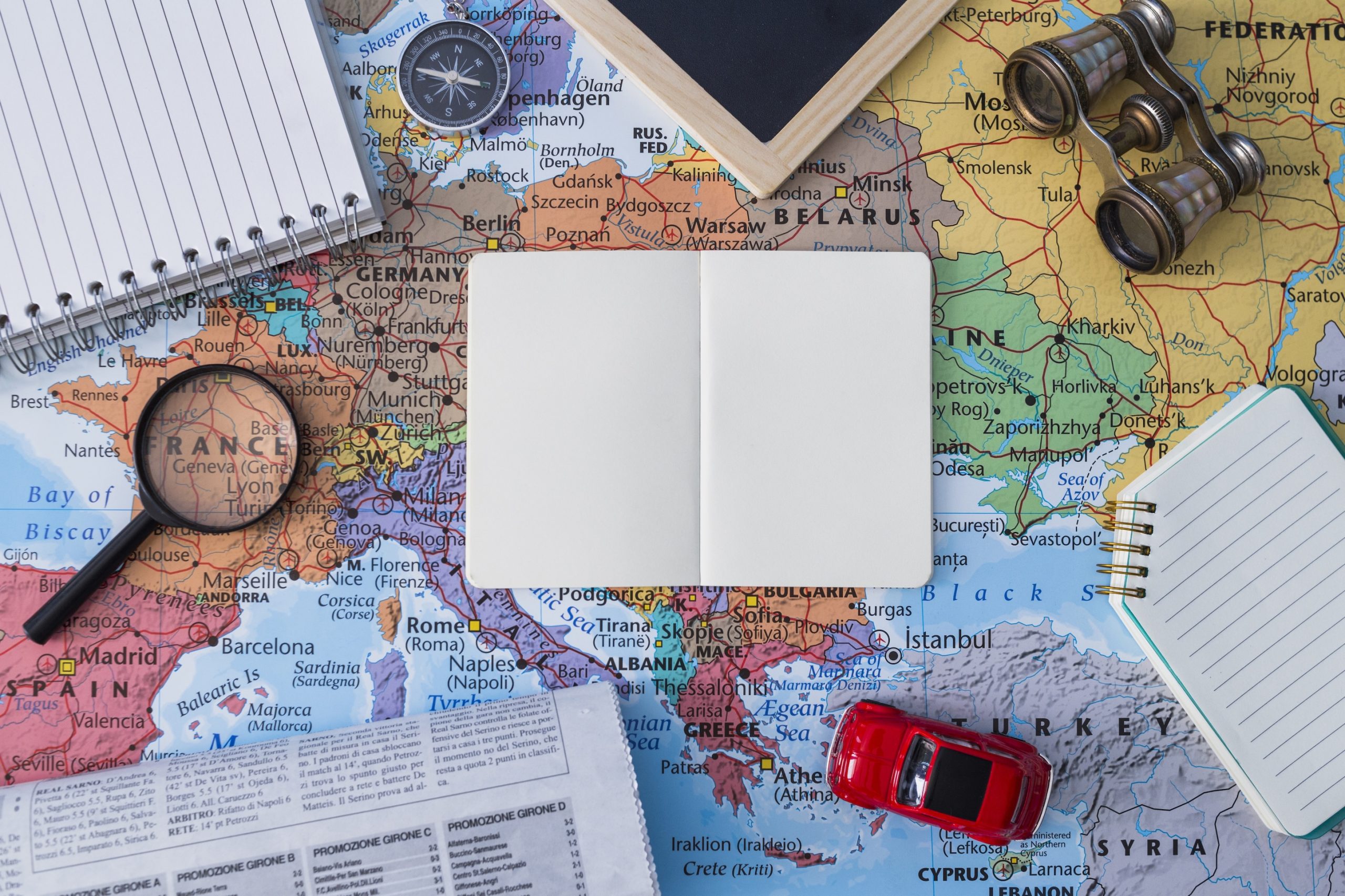 Európa térképén elhelyezett utazási kellékek – nagyító, iránytű, távcső és jegyzetfüzet – egy nyitott üres füzet körül, amely az utazástervezés és felfedezés hangulatát idézi.