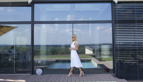 Movik Gabriella ingatlanközvetítő fehér ruhában egy luxusingatlan üvegfala előtt