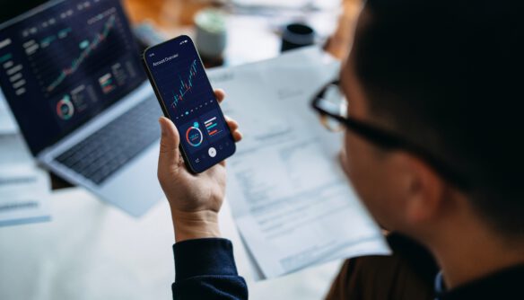 A képen egy íróasztalnál dolgozó személy látható, aki egy okostelefonon megjelenő pénzügyi irányítópultot figyel. A telefon kijelzőjén különböző grafikonok, diagramok és statisztikai mutatók jelennek meg, amelyek a befektetések, a pénzügyi teljesítmény vagy a piaci folyamatok nyomon követésére utalnak. A háttérben egy nyitott laptop látható hasonló grafikonokkal, valamint az asztalon különféle dokumentumok hevernek, ami azt sugallja, hogy a felhasználó több eszközön elemzi az adatokat. A kép az adatalapú döntéshozatalt, a digitális pénzügyi elemzést és a piaci trendek folyamatos követését szimbolizálja.