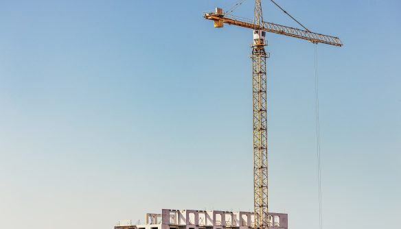 Magas, sárga építési daru magasodik egy épülő, szerkezetkész többemeletes társasház mellett, tiszta, kék égbolt előtt. A kép tökéletesen szemlélteti az új építésű lakások piacát, a lakópark-fejlesztéseket, az építőipari beruházásokat és a lakáskínálat bővülését.