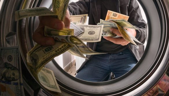 Egy férfi bankjegyeket szór egy mosógép dobjába. A kép a mosógép belsejéből készült, és a pénzmosást szimbolizálja. A PMT-szabályozás változását bemutató cikk illusztrációja.