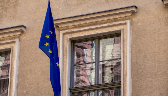 Az Európai Unió kék, sárga csillagos zászlaja függőlegesen kitűzve egy klasszikus, bézs homlokzatú épület ablaka mellett. A fotó tökéletesen illusztrálja az európai uniós szabályozásokat, az EU-s támogatásokat, a nemzetközi gazdasági folyamatokat és a hazai ingatlanpiacot érintő uniós direktívákat.
