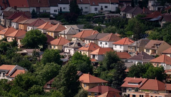 Magasból készült fotó egy sűrűn beépített, domboldali lakóövezetről, piros cseréptetős családi házakkal és kertes ingatlanokkal, zöld fák ölelésében. A kép tökéletesen illusztrálja a kertvárosi ingatlanpiacot, a családi házak kínálatát és a zöldövezeti lakhatási lehetőségeket.
