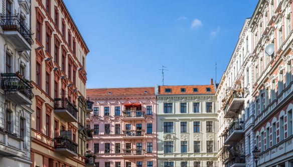 Napfényes belvárosi utcarészlet színes, díszes homlokzatú, többemeletes polgári társasházakkal és erkélyekkel, tiszta kék égbolt alatt. A látványos városi életkép tökéletesen illusztrálja a belvárosi használt lakások piacát, a téglaépítésű polgári bérházak kínálatát és a fővárosi lakókörnyezet hangulatát.